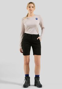 Γυναίκα που στέκεται με ανοιχτόχρωμο μακρύ μανίκι μπλούζα με λογότυπο βουνού, μαύρες βερμούδες, μπλε κάλτσες και μαύρες ορειβατικές μπότες σε γκρι φόντο.