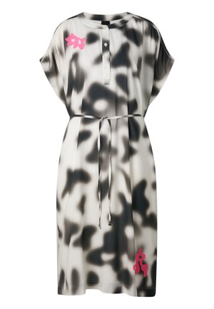 Woman wearing a black and white tie-dye dress with pink floral accents, a waist tie, and black flip-flops, standing with hands in pockets.