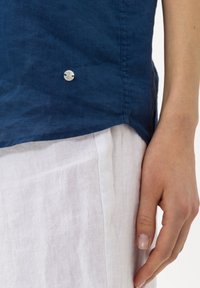 La mano destra poggia accanto a dei pantaloni di lino bianchi e una camicia di lino blu navy con un piccolo bottone decorativo vicino all'orlo.