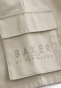 Béžová tkanina s hladkou textúrou, so veľkou kapsou a embosovaným logom "BAKER LONDON BY TED BAKER" v neutrálnych farbách.