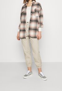 Femme portant un pantalon de jogging beige, un haut blanc, une surchemise à carreaux rose et noir, et des baskets grises et blanches, debout devant un fond blanc.