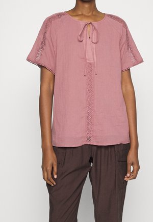 Person trägt eine staubig pinke Kurzarmbluse mit Spitzenbesatz an Ärmeln und Vorderseite, kombiniert mit dunkelbraunen Cargo-Hosen.