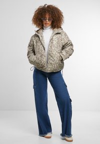 Pufota jaka gaisā bēžā krāsā ar kamuflāžas rakstu, apvienota ar plaša kāju džinsiem, kuriem ir kravas kabatas un plīsušas apakšmala.