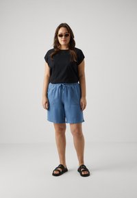 Černý top s krátkým rukávem, světle modré kraťasy s elastickým pasem a kapsami, a černé sandály. Modelka má na sobě sluneční brýle, stojí na neutrálním pozadí.