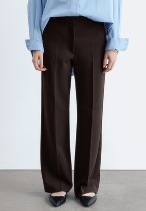 Donkere bruine broek met rechte pijpen, gedragen met een lichtblauw overhemd en zwarte puntige ballerina's op een grijze vloer.