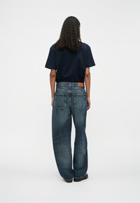 Jeans din denim albastru cu picioare largi și talie medie, prezentând o etichetă din piele maro pe talia din spate și plasamente distincte pentru buzunarele din spate.