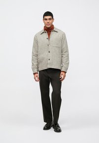 Muž v béžovom károvanom saku na hrdle s hrdzavým rolákom, čiernymi nohavicami a čiernymi topánkami, stojí proti jednoliatemu bielému pozadiu.