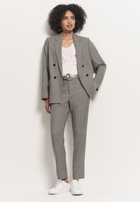 Szary kostium w kratę z podwójnym żakietem i dopasowanymi spodniami, w połączeniu z białą koszulą z grafiką i białymi sneakersami.