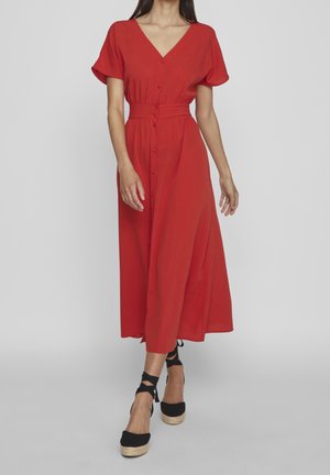 Kobieta ubrana w czerwone midi zapięte na guziki, z krótkimi rękawami oraz czarne espadryle na koturnie z wiązaniami wokół kostek.