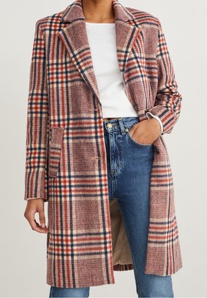 Mujer con un abrigo largo de cuadros en rojo, azul marino y beige sobre una camiseta blanca y vaqueros azules, con la mano en el bolsillo.