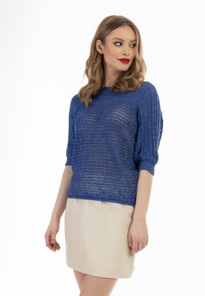 Maglione blu a maglia con un design loose e testurizzato, con maniche corte, indossato su una gonna attillata di colore chiaro.