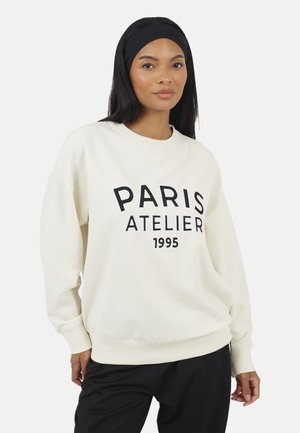 Donna che indossa una felpa bianca con la scritta "PARIS ATELIER 1995", pantaloni neri e una fascia nera, in piedi davanti a uno sfondo semplice.