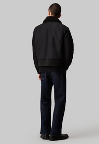 Chaqueta bomber negra con un cuello de piel oscura texturizada y puños acanalados. Se lleva con jeans azul oscuro y zapatos negros, vista desde la parte posterior.