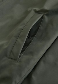 Mørkegrøn stof med en glat tekstur, der har en vertikal lomme med knaplukning. Rene linjer og minimalistisk design.