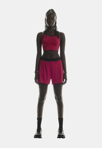 Rød træningscrop top og matchende shorts med en sort talje. Modellen bærer sorte ankelstrømper og sneakers. Glat, tætsiddende stoftekstur.