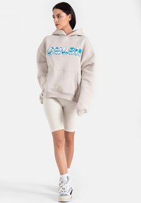 Beżowa bluza z kapturem z niebieskim tekstem graficznym, rybackie białe szorty rowerowe oraz sneakersy z szarymi i czarnymi akcentami.