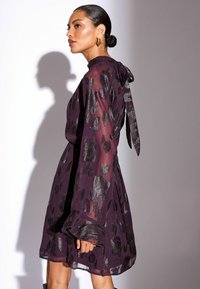 Lila Kleid mit transparenter, langärmliger Gestaltung, floralem Silbermuster, hohem Ausschnitt mit gebundenem Detail im Rücken und einer fließenden Silhouette.