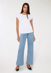 Kobieta stoi w białej koszulce z krótkim rękawem, jasnoniebieskich spodniach o szerokich nogawkach i czarnych szpilkach na wysokim obcasie na jednolitym tle.