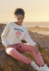 Licht crème sweatshirt met 'Sunrise' grafiek en koraalroze joggers, met een trekkoord met kwastjes; gedragen met witte sneakers.