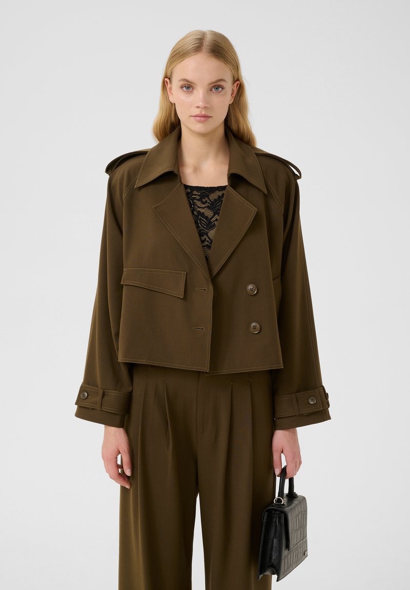 Olijfgroene cropped jas met dubbele knopen, brede revers en subtiele stiksels. Gecombineerd met geplooide broek en een zwarte handtas.