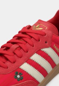 Κόκκινο αθλητικό παπούτσι Adidas Samba με τρεις λωρίδες σε ανοιχτό καφέ χρώμα, ανθικό σχέδιο στο πλάι και υφασμάτινη επιφάνεια με χρυσή σήμανση.