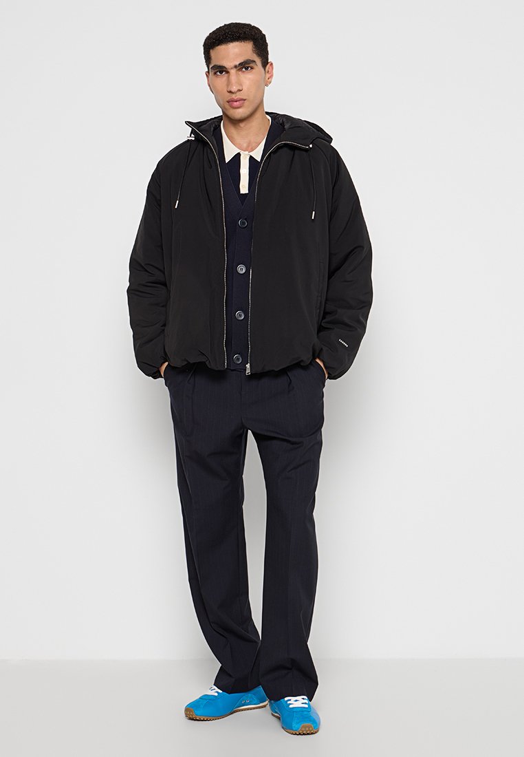 Zwarte capuchon bomberjack, rits aan de voorkant, met trekkoorden. Gelaagd over een marineblauwe cardigan met knopen. Gecombineerd met marineblauwe broek met streepjes en blauwe schoenen.