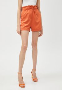 Shorts naranjas de cintura alta con cinturilla fruncida, botones frontales y dobladillos enrollados, combinados con sandalias de tacón alto naranjas con tiras.