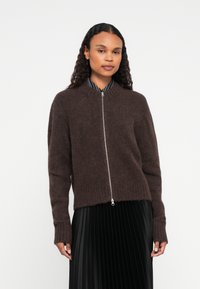 Γυναίκα που φοράει ένα καφέ φούτερ με φερμουάρ και γούνινες λεπτομέρειες πάνω από μια μαύρη πλισέ φούστα, που στέκεται μπροστά σε έναν απλό λευκό τοίχο.