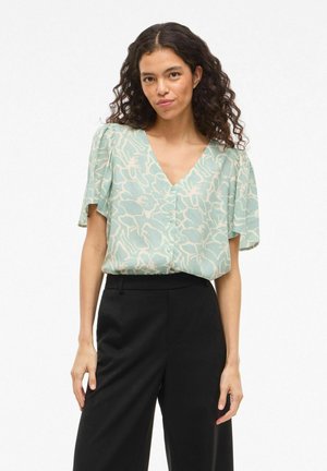 Femme aux cheveux bouclés portant un chemisier à col en V vert menthe à motifs floraux et un pantalon noir taille haute, debout devant un fond blanc.