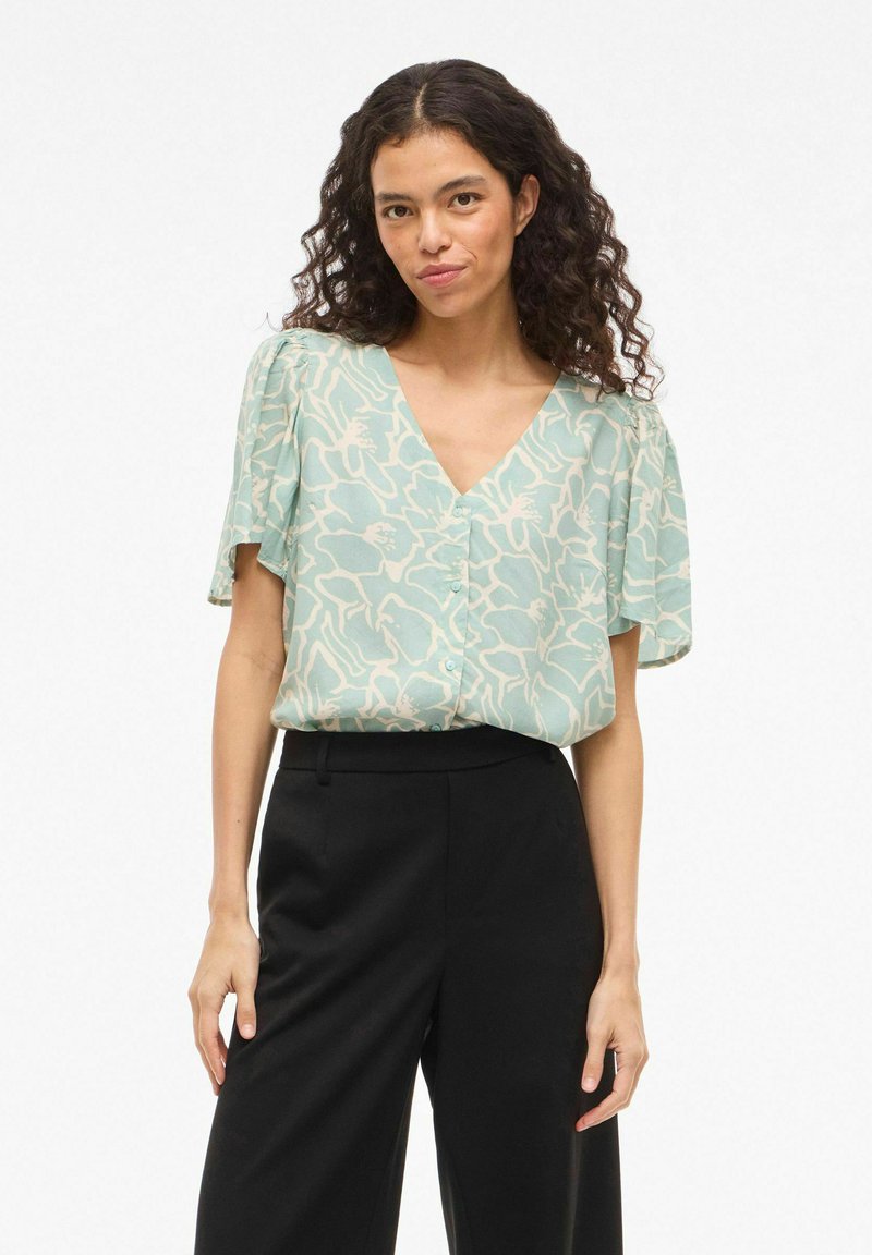 Donna con capelli ricci che indossa una blusa a fiori verde menta con scollo a V e pantaloni neri a vita alta, in piedi contro uno sfondo bianco.