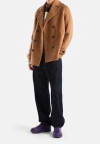 Abrigo de lana marrón de doble botonadura con grandes botones, combinado con pantalones de mezclilla azul oscuro y zuecos morados. Color sólido, diseño sencillo.
