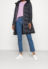 Schwarzer gesteppter Daunenmantel mit Kapuze, vertikalen Steppmuster, kombiniert mit einem hellvioletten Pullover, blauen Jeans mit geradem Bein und weißen Sneakern.