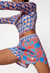 Pitkähihainen, vartalonmyötäinen paita, jossa on geometrinen kuvio sinisessä, oranssissa ja valkoisessa. Vastaavat shortsit rohkealla kukkakuviolla punaisessa ja sinisessä.