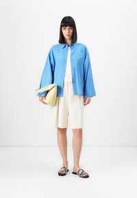 Woman stands wearing a loose blue button-up shirt, white top, cream knee-length shorts, snakeskin sandals, and holding a pale yellow handbag.