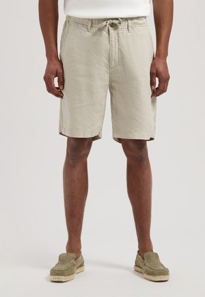 Man draagt beige shorts met trekkoord en olijfgroene instapschoenen, staand tegen een effen achtergrond.