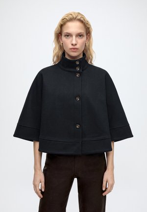 Femme blonde portant une veste style cape noire boutonnée avec des manches larges et un col montant, debout devant un fond uni.
