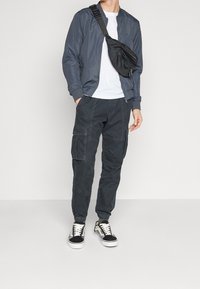 Donkergrijze bomberjack, wit T-shirt, cargobroek met zakken, zwarte sneakers met witte veters en een zwarte schoudertas.