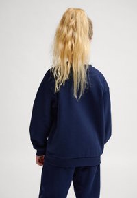 Marinblå oversized sweatshirt, tillverkad i mjukt tyg, med ribbade ärmslut och nederkant, sedd bakifrån. Långt, vågigt blont hår synligt.