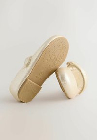 Gold glitter ballet shoes with an elastic strap, rounded toe, and a textured rubber sole featuring a ballet motif.