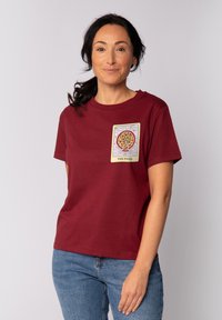 Bordeauxrotes Baumwoll-T-Shirt mit kurzen Ärmeln, das auf der Vorderseite ein bedrucktes Pizzamotiv in rechteckigem Design aufweist. Darunter werden blaue Jeans getragen.