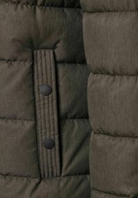 Detalje af mørke olivengrøn dynejakke med ribstrikket stofaccent, sort knap med trykknap og quiltet tekstur, der viser syede sektioner.