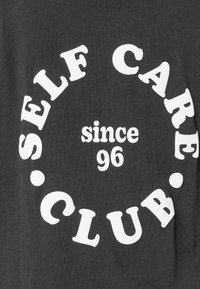 Dunkler Stoff mit weißem Text in kreisförmiger Anordnung, der "SELF CARE CLUB" und "seit 96" in der Mitte des Kreises liest.