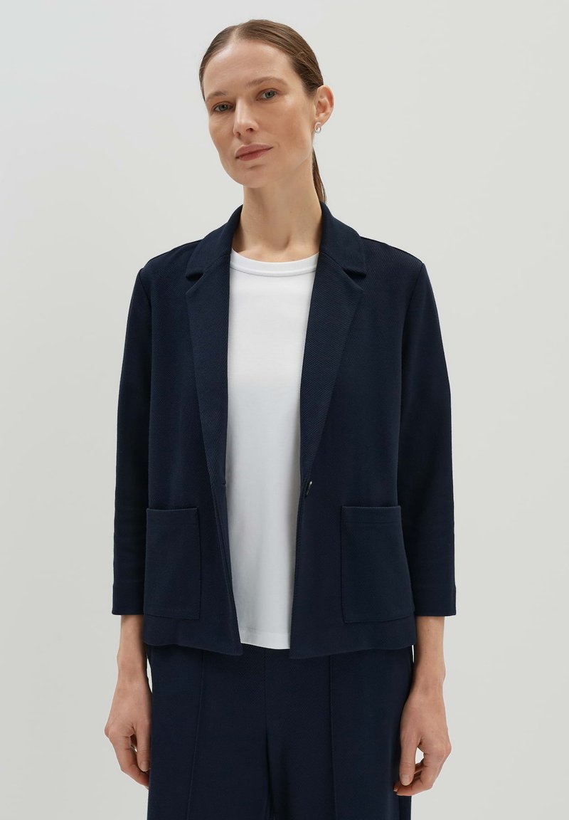 Marineblazer aus strukturiertem Stoff mit Reverskragen, zwei Fronttaschen und dreiviertel Ärmeln, getragen über einem weißen Oberteil.