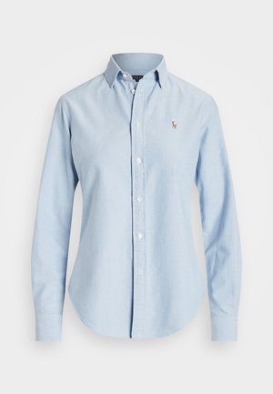 Camisa azul clara de manga larga, confeccionada en algodón, con cuello puntiagudo y un pequeño logo bordado en el lado izquierdo del pecho.