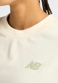 Close-up van de nek en bovenste borst in een lichtbeige shirt met een lichtgroen New Balance-logo aan de linkerkant.