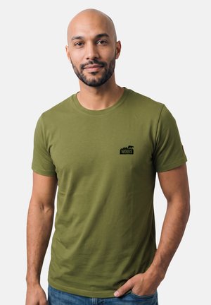 Mann mit Bart trägt ein olivgrünes kurzärmeliges T-Shirt mit einem kleinen schwarzen "Works"-Logo auf der Brust und blaue Jeans.