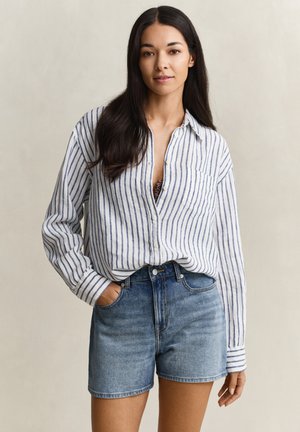 Femme aux longs cheveux bruns portant une chemise blanche à rayures bleues boutonnée et rentrée dans un short en denim taille haute, sur fond uni.