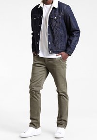Dark denim jacket with white fleece collar, white t-shirt, olive green pants, and white sneakers, standing against a light background.