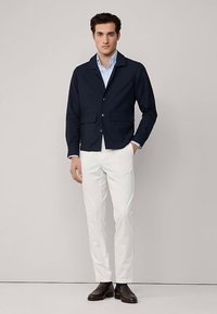 Marineblauwe katoenen jas met kraag, twee zakken aan de voorkant en knoopsluiting, gecombineerd met witte broek en donkerbruine schoenen.