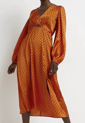 Nainen pukeutuneena oranssiin satiini-midi-mekkoon, jossa mustia pilkkuja, pitkät paisutetut hihat, etutasku ja sivuhalkio.